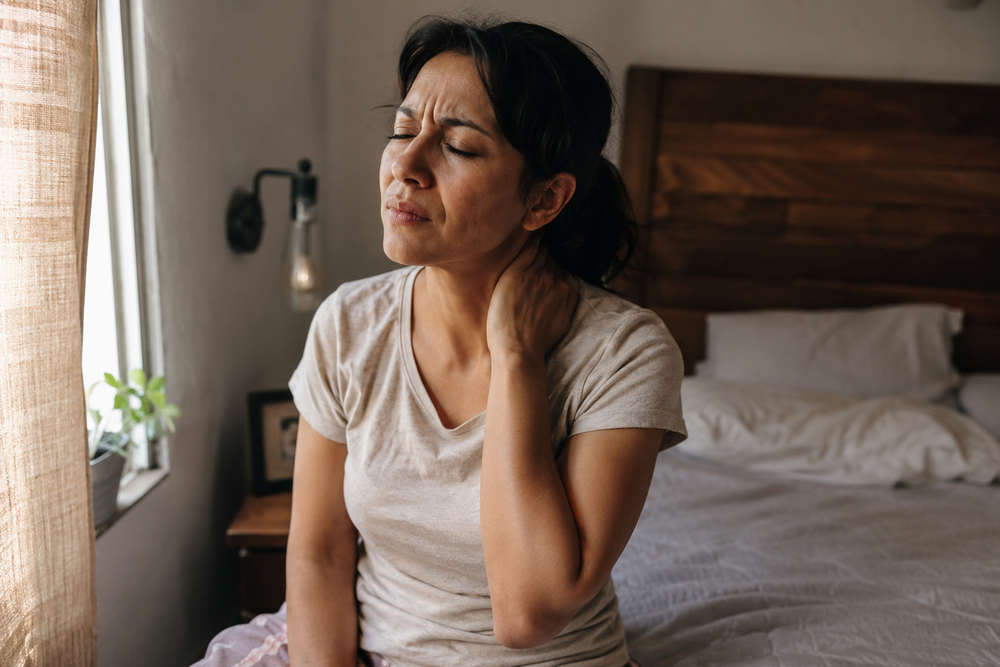Fotografía realista y emotiva de una mujer mexicana de unos 35 años sentada en el borde de la cama por la mañana, con una mano en el cuello y expresión de dolor y agotamiento, luz natural suave entrando por una ventana, ambiente íntimo de recámara real, tonos cálidos y ligeramente desaturados, estilo documental/editorial, foco en la emoción del rostro y el gesto de la mano en el cuello, sin texto ni marcas de agua, alta resolución