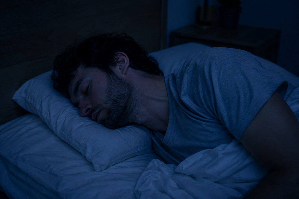 Fotografía nocturna sutil de una persona durmiendo de lado con el cuello en un ángulo forzado e incómodo sobre una almohada plana, luz azulada tenue de madrugada, habitación en penumbra, se percibe la tensión en el cuello aunque la persona esté profundamente dormida, estilo editorial/documental, tonos fríos, sin texto ni marcas de agua