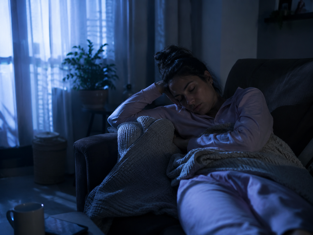 Fotografía realista y emotiva de una mujer en pijama durmiendo incómoda en un sillón, envuelta en una cobija pequeña, luz tenue de madrugada entrando por una ventana, ambiente silencioso y solitario, tonos fríos, estilo documental/editorial, sin texto ni marcas de agua