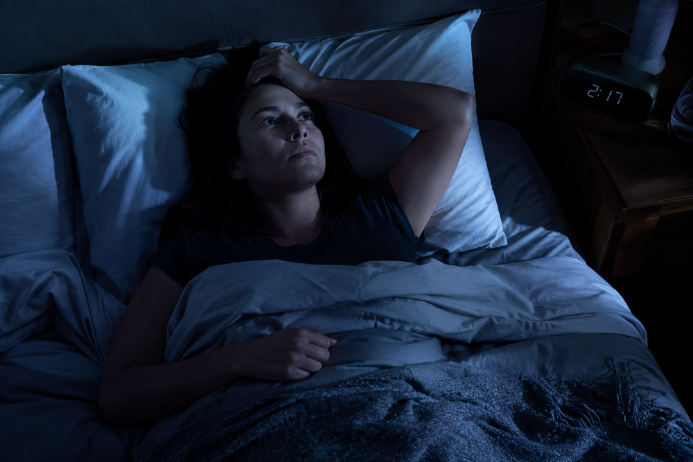 Fotografía realista y emotiva de una mujer despierta en la cama de noche, mirando el techo con expresión de frustración y agotamiento, luz tenue de luna entrando por la ventana, el reloj en el buró marcando las 2:17 AM visible pero no protagonista, ambiente íntimo de recámara real, tonos azulados fríos, estilo documental/editorial, sin texto ni marcas de agua
